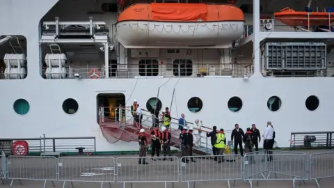 EPA/Shutterstock People walking off a ramp off the ship surrounded by workers