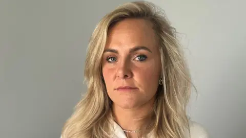 BBC Hannah Farrell, a nurse, looks into the camera. She has a neutral expression on her face. We can see her from the shoulders up. She has shoulder-length blonde hair and is wearing a white or cream top. She has a metal chain around her neck. She is standing in a room, there is a plain grey-coloured wall behind her.