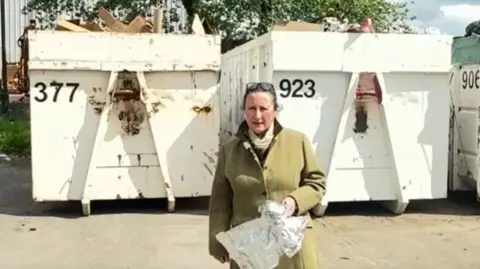 Councillor Elissa Swinglehurst wearing a green tweed coat holds up rubbish in both hands while standing in front of white recycling bins.