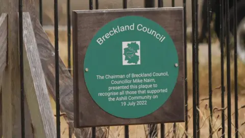 A green plaque reads: Breckland Council. The chairman of Breckland Council, Councillor Mike Nairn, presented this plaque to recognise all those that supported the Ashill Community on 19 July 2022