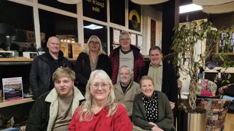 Deana Bamford Eight board members of Coalville CAN are gathered round in their HQ at night time in their coats and jumpers, sitting looking directly at the camera