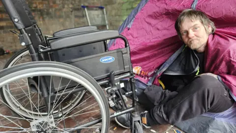 Aisha Iqbal/BBC A man in a tent sat next to a wheelchair in Bradford