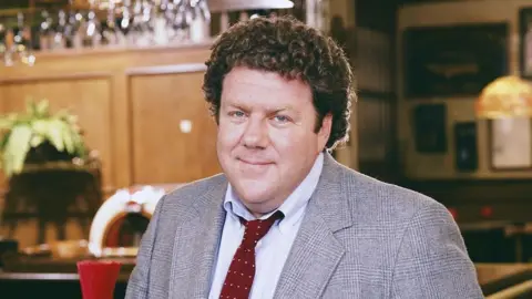 George Wendt as the character Norm wearing a jacket and red tie