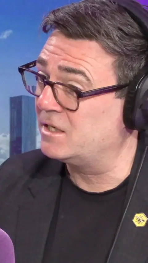 Andy Burnham, a man with black hair and glasses and black t shirts and a blazer wearing a bee badge