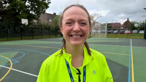 Sarah Dodd is smiling as she stands on an outdoor pitch. She is wearing a hig visibility long-sleeved top and a blue lanyard. Her straight brown hair is tied back.