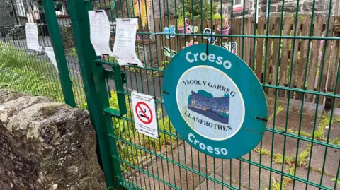 The closed gate of Ysgol Y Garreg, a modern, green wire gate set into a fence of the same type. A modern notice with the school name on - slightly worn - and a no smoking sign are attached to the gate by cable ties