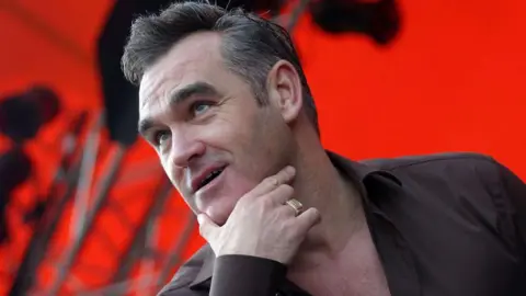 Head and shoulders of Morrissey on stage with his right hand thumb and index finger cupping his chin. He is wearing a gold signet ring, a black shirt and the background is lit red with scafolding
