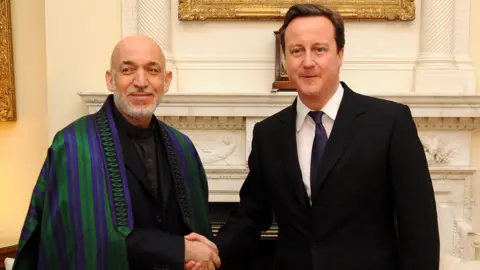 Getty Images A meeting at Downing Street in 2011 between UK Prime Minister David Cameron, wearing a dark suit and tie, and Afghan President Hamid Karzai, who is wearing a dark suit and shirt and an embroidered chapan, an Afghan coat or cape, with purple and green stripes. They are shaking hands as they stand in front of a marble fireplace and paintings in gilded frames