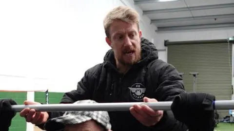 Patrycja Boryka/BBC A man with brown short hair swept to the side is holding a long weight and looking to the side at someone's hand which is holding it either side with black gloves on. His mouth is partially open as he assists the person, whose face cannot be seen but is wearing a grey and black cap, who is lifting the weight.