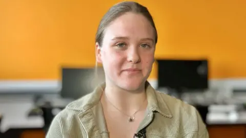 Martin Giles/BBC Eve in a computer room. The background, which shows an orange wall and two computer monitors, is out of focus. Eve has brown hair tied back. She is wearing a beige corduroy jacket. She is wearing a silver pendant.