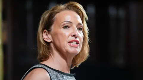 PA Media Reform MP Sarah Pochin wearing a sleeveless grey textured top, standing indoors against a dark blurred background with soft lighting