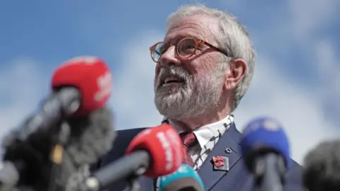 Gerry Adams, a man with grey hair and beard in a navy suit jacket, smiling in front of numerous microphones