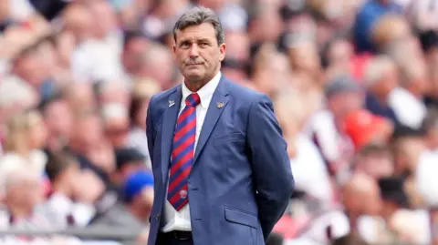 PA Media Media is a man with short white hair, blue and red striped tie, blue blazer and a white shirt, standing behind him on a football field. The crowd blurred in the stands behind him.
