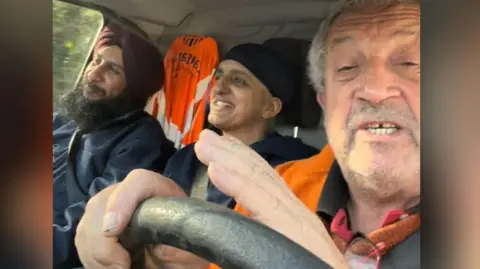 Marcus Lapsa Bill Mato, Kully Kaur Deol and Marcus Lapsa are smiling in their van driving down to Pontypridd. They are wearing blue jackets and Marcus Lapsa is wearing an orange hi-vis jacket. Mr Lapsa is driving.