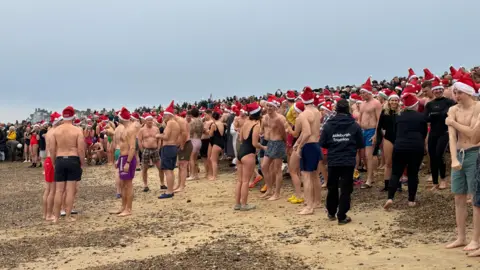 Swimmers on the beach preparing to take to the sea for a Boxing Day dip in Aldeburgh at Moot Hall. People are topless or in swimsuits and are wearing red and white Santa hats.