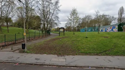 Google The entrance to a park - trees are on the grass behind a green fence on the left. A noticeboard is a short way up the footpath next to a grassy area in the centre. A skate park is visible on the right.