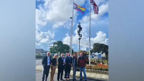 Great Yarmouth Borough Council The Progress Pride flag being flown by Great Yarmouth Borough Council. Councillors are stood underneath the flag smiling at the camera. There is another flagpole with a union jack flag flying from it.