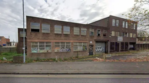 Google A two-storey building, seen from the outside and with graffiti on the downstairs windows, is most prominent in the photo. The pavement and part of a road is in the foreground.