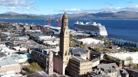 A coastal cityscape with a tall clock tower in the foreground, surrounded by historic and modern buildings. Behind it, a large white cruise ship is docked beside red industrial cranes. Beyond the harbour, green hills and mountains rise under a partly cloudy blue sky.