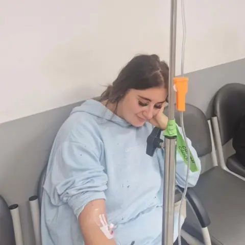 Paige Roberts Paige Roberts sitting down in a hospital chair having IV fluids administered. She is wearing a light blue hospital gown and is looking down at the floor 