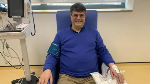 A man with glasses and blue sitting in a chair in hospital with a cannula inserted in his hand. 