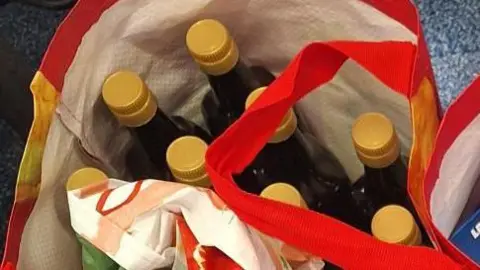 A large collection of items in several large bags for life. You can see packets of coffee, chocolate bars, olive oil bottles and toiletries. They are all placed on the ground on top of a grey carpet. 