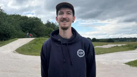 Simon Thake/BBC A tall man in a baseball cap and blue hoodie stands on an empty BMX track.