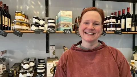 BBC/Julie Mariotti A woman smiling in front of a shelf full of products in a delicatessen. 