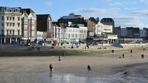Properties by a seafront. There are people on the beach, some walking their dogs.