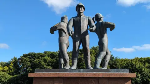 Alamy A blue sky and a horizon of green trees in full bloom. In front, a bronze statue of three oil workers in boiler suits and helmets, back-to-back and facing three directions. They are on a red granite plinth.