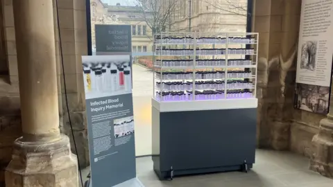 Photograph of the memorial to the victims of the NHS blood infection scandal. The image shows a display case, with blood vials containing personal messages from those affected.