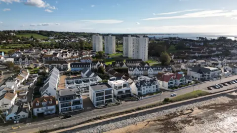 Aerial view of a coastal residential area with a mix of modern apartment buildings, townhouses and detached homes. A seafront road runs alongside a stone sea wall and a narrow beach in the foreground. Three tall white tower blocks stand among lower-rise housing, with green fields and rolling countryside behind. The sea stretches across the horizon under a bright blue sky with scattered clouds.