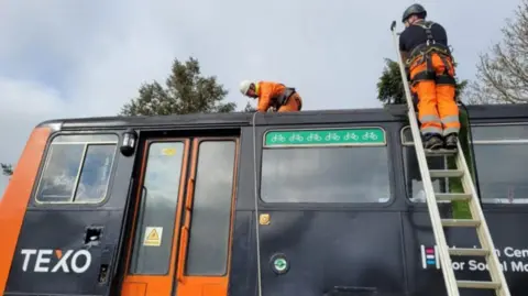Northern Two engineers wearing orange and black work clothing and harnesses are repurposing the driver's cab of the Pacer train. One stands atop of a ladder, while the other fixes something on the roof of the train. The train is branded 'TEXO' and is orange and black.