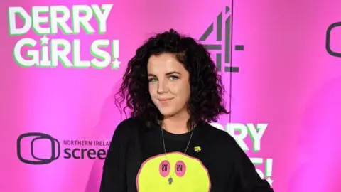 Getty Jamie Lee- O'Donnell has long brown bushy hair and is wearing a black T-shirt with a yellow smiley face. She is standing in front of a pink board with a number of signs. 
