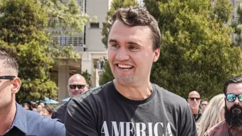 Getty Images Charlie Kirk stands outside in a black and white T-shirt smiling and surrounded by people in May 2025.