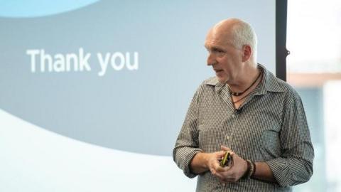Mark Foulkes, standing in front of a screen delivering a presentation. He is a bald white man with fine grey hair around the edge of his head and he can be seen wearing a checked shirt with the sleeves rolled-up