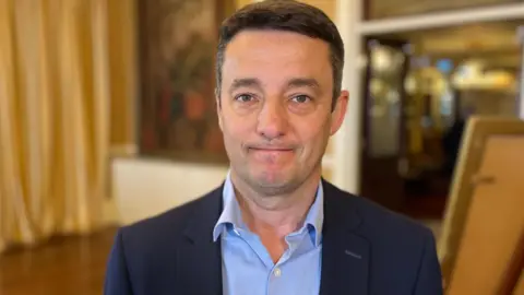 Boley Smillie is pictured. He has black hair and brown eyes, and is wearing a blue blazer with a blue shirt. 