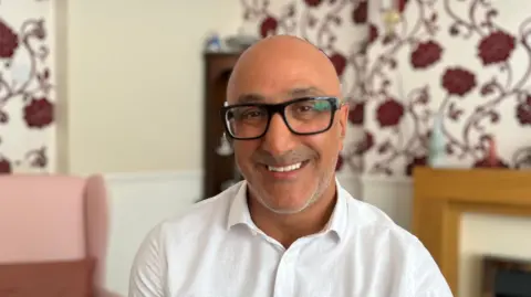 A middle-aged man with thick black-rimmed glasses is smiling at the camera, wearing a white shirt and sitting in a living room.