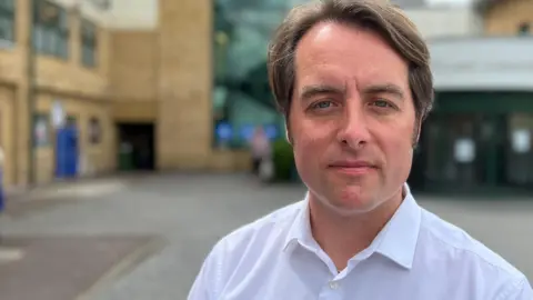 SIMON DEDMAN/BBC Thom Lafferty stands outside the newer front entrance of the Princess Alexandra Hospital in Harlow and looks down the barrel of the camera. It has beige brick and a glass staircase. He is wearing a white collared shirt and has blue eyes and brown hair