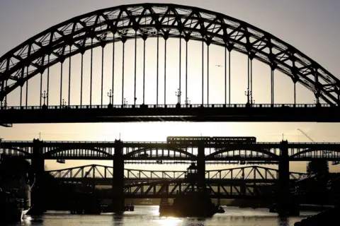 Owen Humphreys/PA Media As the sun sets behind the bridges, they are cast in silhouette. The Tyne Bridge, closest, is arched and made in a metal criss-cross pattern. A train crosses the rail bridge beyond. And there are more bridges beyond that. The sun is reflected in the water below.