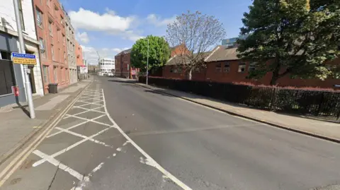 Google A Google street view photo of Glasshouse Street. It's a road in the city with tall buildings either side, lamp posts and a couple of trees on the right.