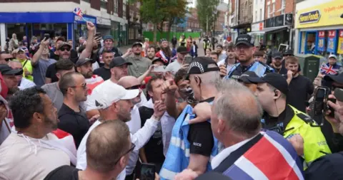 PA Media A crowd of people, some wearing flags around their shoulders, square up to police who are engaging with protesters in Nuneaton. In the background, shops can be seen on both sides of the street.