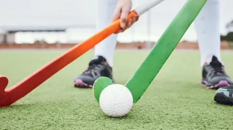 A close up image showing two people playing hockey on an astro-turf pitch. The person in the background is holding an orange stick while the person in the foreground with a green stick prepares to hit the ball. 