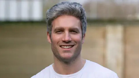 Michael James Close up of Dafydd smiling looking at the camera. He is standing in front of a brick wall but the background is blurred. He wears a white t-shirt and has grey hair.