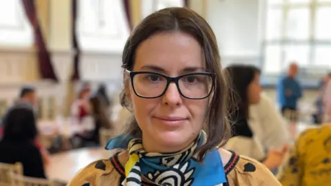 Jonny Michel/BBC A woman with dark hair and glasses wearing a colourful blue pattered scarf.