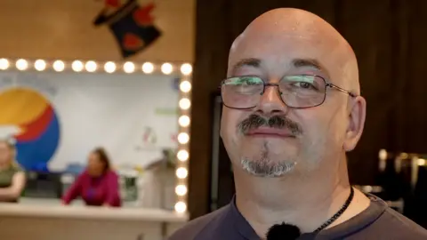 Jamie Niblock/BBC Jonathan Conlin looking down the camera, standing in the school uniform exchange shop.