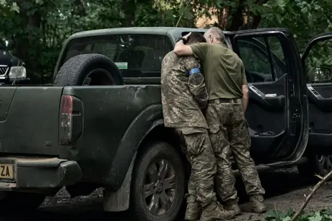 Kostiantyn Liberov/Libkos via Getty Images Two soldiers embrace, with one resting his head on the other's shoulder, both dressed in military outfits and standing next to a truck with its doors open and with bullet marks on its side, in the north-eastern Sumy region of Ukraine on 14 August 2024.