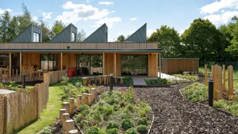 Daniel Hopkinson A garden with a bark path and wooden planters, with a wooden triangular building in the background. There are also trees in the background and small plants in the garden.