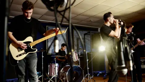 Jamie Niblock/BBC All band members are dressed in black. Luke, the guitarist is holding his white and black electric guitar. Joe, the drummer, is in the backdrop holding his sticks. Front man Ben is grasping the microphone, whilst bassist Ed is clutching his bass guitar.