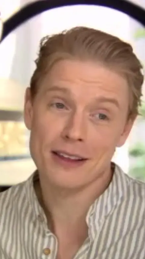 Freddie Fox has blonde hair swept back, he is wearing a grey and white striped collared shirt. 